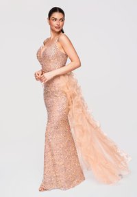 Femme portant une robe à sequins pêche scintillante avec de fines bretelles et une longue traîne en tulle froncée, posant avec les mains jointes et regardant sur le côté.