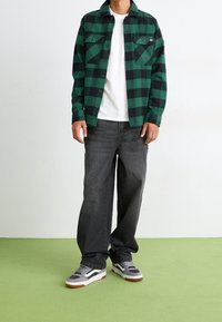 Chemise en flanelle à carreaux verts et noirs, t-shirt blanc en dessous, pantalon en denim gris, baskets gris et blanc avec des accents rouges.