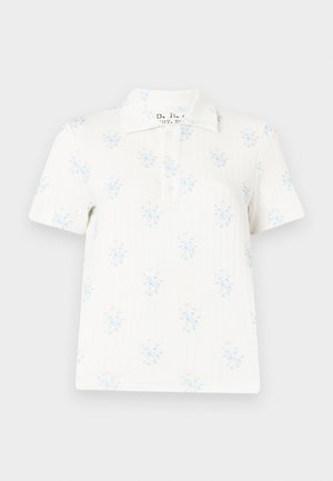 Femme debout les mains dans les poches, portant une chemise blanche à manches courtes, un pantalon marron large à ceinture, et des chaussures blanches pointues.