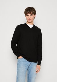 Schwarzer V-Ausschnitt-Pullover aus weichem Material, mit geripptem Muster, langen Ärmeln und tailliertem Design. Trägt ihn über einem weißen Hemd mit blauen Jeans.