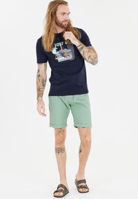 Merihenkinen t-paita painetulla grafiikalla, vihreät shortsit, ruskeat sandaalit. T-paidassa on kuvasto palmupuiden ja surfing-elementtien kanssa.