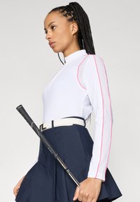 Valkoinen pitkähihaiset paita vaaleanpunaisilla yksityiskohdilla ja verkkomaisella yksityiskohdalla, yhdistettynä laivastonsinisiin hameeseen. Golfmaila nojaa vyötärölle, kiinnitettynä valkoisella vyöllä.