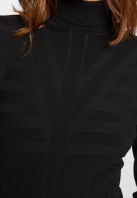 Suéter negro de punto acanalado de cuello alto con patrones geométricos texturizados en el pecho y los hombros, llevado por una persona con cabello castaño rojizo.