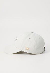 Hvit bomull baseball cap med buet skjerm, teksturert stoff, brodert logo i svart og gull, og en justerbar stropp bak.