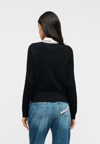 Μαύρο πουλόβερ με μακριά μανίκια και ριμπ τελείωμα, με λευκό γιακά που φέρει την επιγραφή "Bianchi." Φορεμένο πάνω από ανοιχτόχρωμα μπλε τζιν με ξεθωριασμένες λεπτομέρειες.