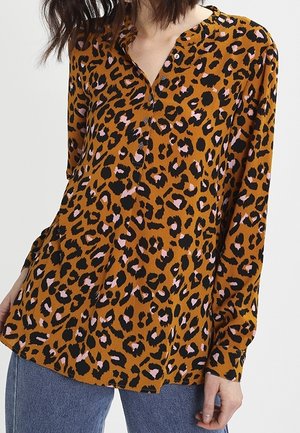 Kvinde iført en brun bluse med lange ærmer, knapper og sort-rosa leopardprint, kombineret med blå jeans.