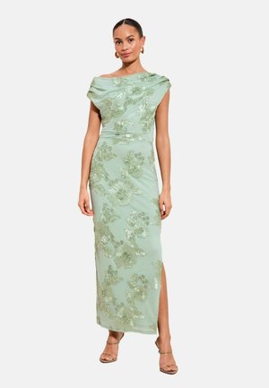 Žena oblečená v svetlozelenej, bezrukávovej, dlhej šatách po zem s kvetinovou výšivkou a bočným rozparkom, doplnená béžovými sandálmi na remienky.