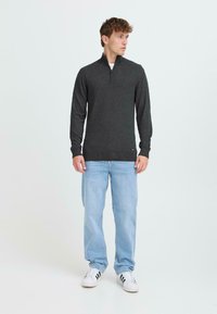Suéter de cuello con cremallera de color gris oscuro en material suave, de manga larga y diseño entallado. Combinado con jeans de color azul claro y zapatillas blancas con rayas negras.