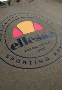 Tissu vert avec le logo ellesse présentant des demi-cercles orange et rose, texte « Authentic Italian Sporting Apparel Established 1959 » en cercle.