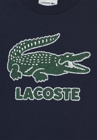Mörkblå bomullströja med en sprucken grön krokodilgrafik och ordet "LACOSTE" i en matchande stil nedanför designen.