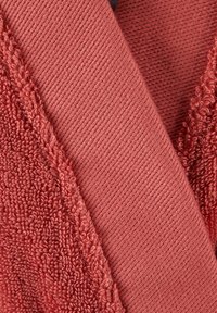 Un tissu couleur rouille présentant deux textures : un tissage en éponge duveteux et une surface lisse et côtelée, créant un contraste entre les motifs et les finitions.