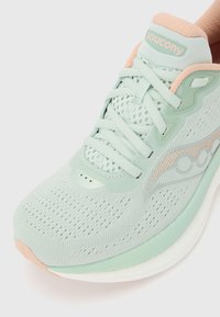 Ανοιχτό πράσινο παπούτσι τρεξίματος Saucony με επάνω μέρος από δίχτυ, λεπτομέρειες σε παστέλ ροδακινί και λευκή μαξιλαρωμένη σόλα, εμφανιζόμενο από κοντινή λοξή κορυφαία όψη.