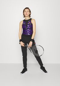 Lila, mit Pailletten besetztes Sportoberteil mit schwarzen Akzenten, kombiniert mit schwarzen Hosen und Handgelenksbändern. Ein Tennisschläger in der linken Hand gehalten.