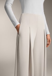 Lichtgrijze wijde broek met een gemêleerde stof, voorzien van voorplooien en zijzakken. Gecombineerd met een lichtblauw shirt met lange mouwen.