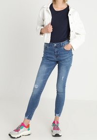 Jeans ajustados azules con detalles desgastados, combinados con una chaqueta blanca corta y una blusa azul marino. Los zapatos tienen acentos de color brillante.