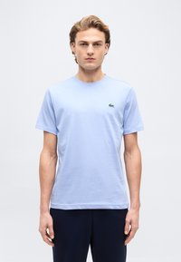 Νεαρός άνδρας που στέκεται με το σώμα στραμμένο προς τα εμπρός, φοράει ένα ανοιχτό μπλε κοντομάνικο μπλουζάκι με ένα μικρό πράσινο λογότυπο κροκοδείλου στο στήθος.