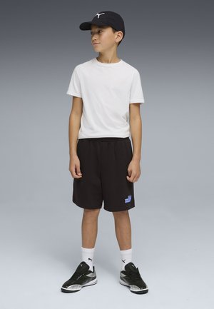 Niño con camiseta blanca, pantalones cortos negros de Puma, gorra negra de Puma, calcetines blancos de Puma y zapatillas negras de Puma, de pie contra un fondo gris.