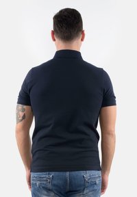 Polo shirt blu navy con colletto corto, design aderente e tessuto strutturato. Maniche risvoltate; presenta un tatuaggio visibile sul braccio.