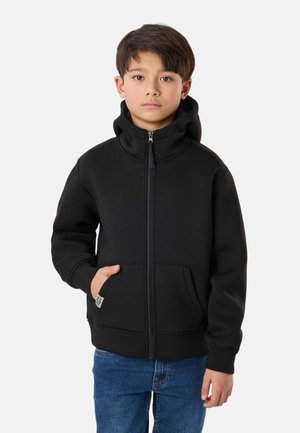 Dreng iført en sort hættetrøje med lynlås og hætten oppe, hånden i lommen, og blå jeans, stående mod en ensartet lys baggrund.