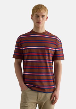 Junger Mann mit kurzen blonden Haaren, der ein kurzärmeliges T-Shirt mit braunen, blauen, roten und weißen Streifen und beigefarbene Hosen trägt, steht mit einer Hand in der Tasche.