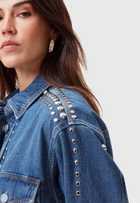 Giacca in denim con borchie metalliche lungo le spalle e le tasche, dal taglio classico e lavaggio blu. La vista ravvicinata mostra i dettagli delle cuciture.