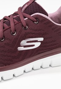 Chaussure de sport bordeaux avec une tige en mesh respirant, semelle extérieure en caoutchouc blanc et accent de logo. Présente des lacets plats et des détails texturés.