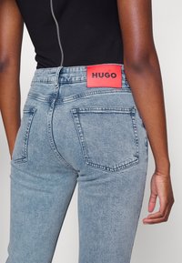 Vista trasera de una persona que lleva unos jeans de mezclilla azul claro con una etiqueta roja "HUGO" y una camiseta sin mangas negra con una cremallera plateada visible.