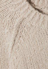 Close-up van beige gebreide sweaterstof met geribde kraag en gedetailleerde textuursteekpatronen.