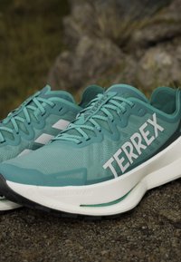 Par turkis Adidas Terrex trail løbesko med hvide mellemsåler på udendørs stenede underlag.