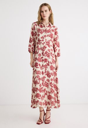 Vrouw draagt een lange crèmekleurige jurk met rood bloemenpatroon, knoopsluiting aan de voorkant, driekwart mouwen en rode high-heeled sandalen met bandjes.