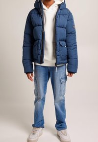 Mörkblå puffjacka med huva, zippered front och två framfickor; bärs över en vit hoodie och matchas med ljusblå cargo jeans och vita sneakers.