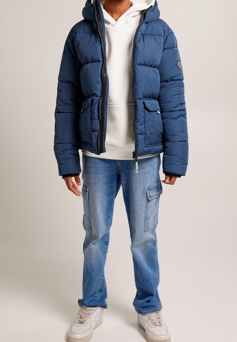 Mörkblå puffjacka med huva, zippered front och två framfickor; bärs över en vit hoodie och matchas med ljusblå cargo jeans och vita sneakers.