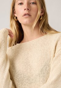 Γυναίκα με ανοιχτόξανθα μαλλιά που φοράει χρυσούς κρίκους στα αυτιά και εκρού πλεκτό πουλόβερ, με ουδέτερο ανοιχτό γκρι φόντο.