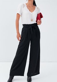 Zwarte wijde broek met plooien en een riem in de taille, gecombineerd met een witte top met kanten accenten. Rode clutch en zwarte laarzen inbegrepen.