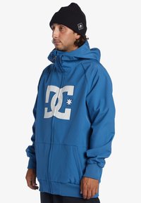 Veste zippée bleue avec capuche, arborant un grand logo blanc à l'avant et des poignets côtelés. Fabriquée en tissu lisse et léger.
