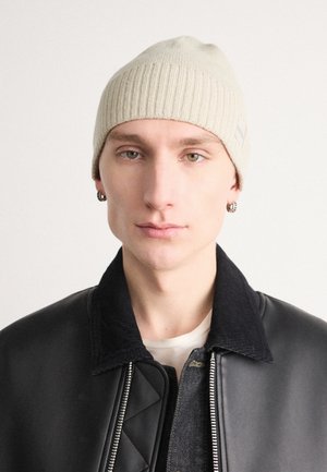 Beige ribatud kudumine beanie sildiga, kantud musta nahast bomber-jakiga, näidates sujuvat tekstuuri ja sobivat disaini.