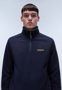 Marineblaue Fleece-Pullover mit einem Halb-Anzip-Kragen, der eine strukturierte Oberfläche und ein Markenlogo auf der Brust aufweist.