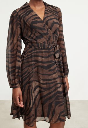 Kobieta ubrana w brązowo-czarną sukienkę kopertową z długim rękawem w paski przypominające zebrę, z przewiązanym paskiem w talii, stojąca na tle jednolitego tła.
