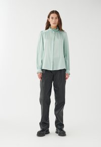 Mintgrün, langärmlige Bluse mit Rüschenausschnitt und gerafften Schultern, kombiniert mit locker sitzenden schwarzen Hosen und klobigen schwarzen Schuhen.