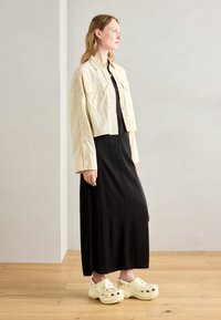 Cropped beige jacket over a black dress, featuring long sleeves and two front pockets. Worn with yellow clogs adorned with decorations.