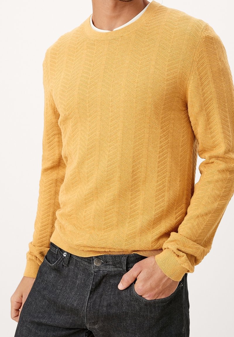 Pull en maille jaune moutarde avec un motif en chevrons texturé, col rond et poignets côtelés, porté sur un jean en denim foncé.