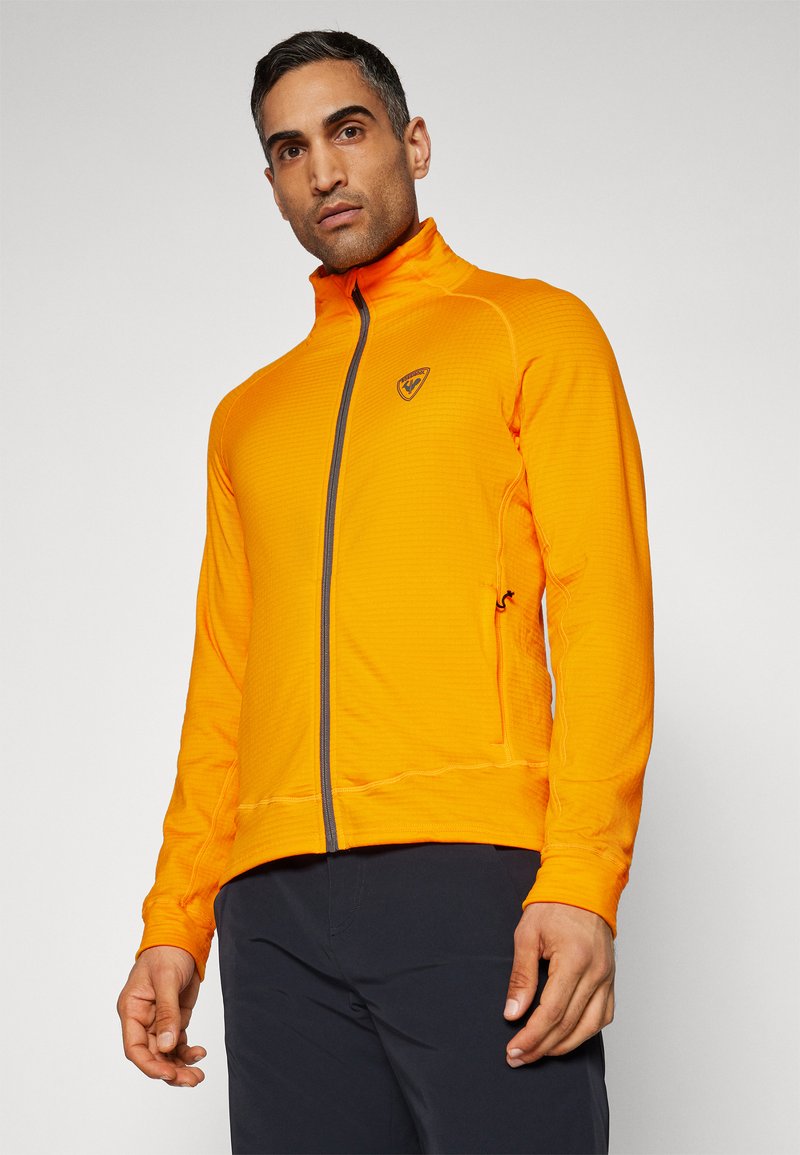 Oranje ritsjack gemaakt van textuurstof, met een staande kraag en een logo op de borst. Rits loopt van boven naar beneden aan de voorkant.