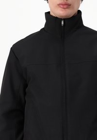 Sort softshell jakke med høj krave, frontlynlås og syninger. Stoffet har en glat tekstur og er letvægtigt.