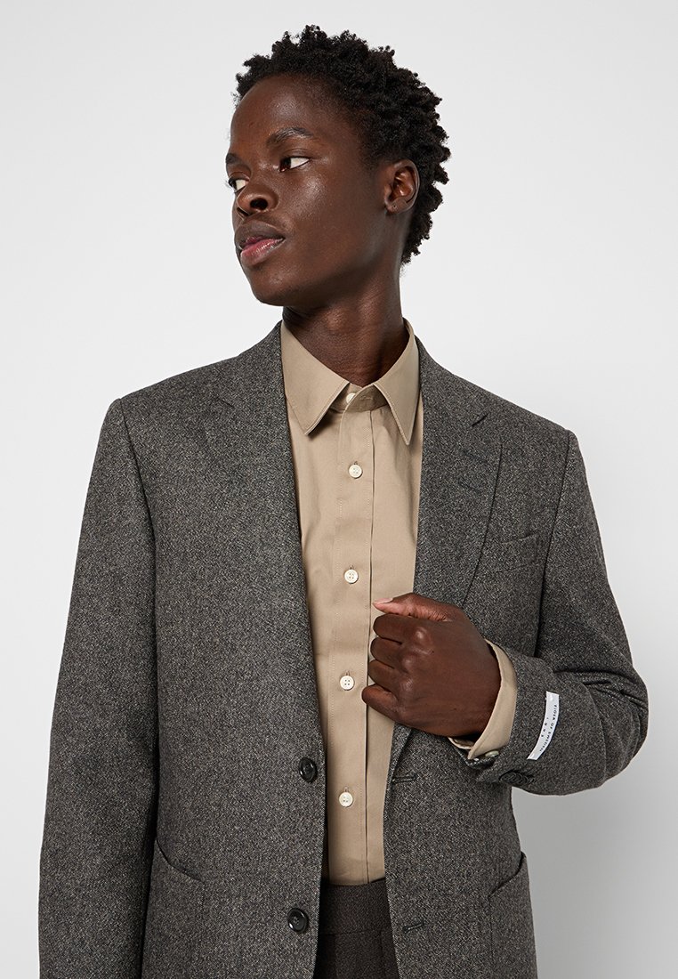 Junge Person mit kurzen lockigen Haaren, die ein graues Tweed-Blazer über einem beigen Hemd mit Knopf trägt und mit neutralem Gesichtsausdruck zur Seite schaut.