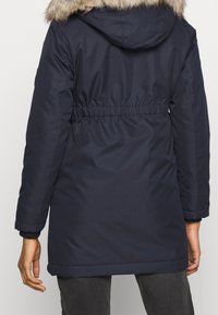 Laivastonsininen toppatakki, jossa on turkisreunainen huppu, joustava vyötärö ja resorit hihansuissa. Pehmeä rakenne, käytännöllinen muotoilu kylmään säähän.