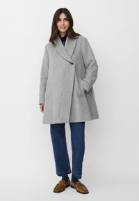 Kvinna stående iförd en grå oversized kappa, blå jeans, marinblå topp och bruna loafers, leende med en hand i fickan på sin kappa.