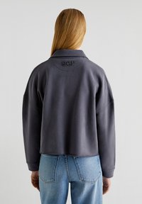 Beskarvet grå sweatshirt med krave, med broderet "SGP" på ryggen og lange ærmer. Sammen med lyseblå denimjeans.