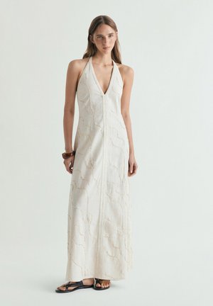 Mujer con vestido maxi blanco de cuello halter con sutiles patrones en espiral, sandalias negras y una pulsera marrón, de pie contra un fondo liso.