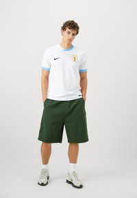 Hvid Nike trøje med lyseblå detaljer og Uruguay-emblem, parret med grønne shorts. Model iført grå sneakers og hvide sokker.
