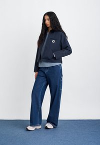 Ναυτικό σακάκι με φερμουάρ τσέπη, φορεμένο πάνω από γκρι τοπ, συνδυασμένο με φαρδιές τζιν παντελόνες και λευκά αθλητικά παπούτσια σε ανοιχτό φόντο.